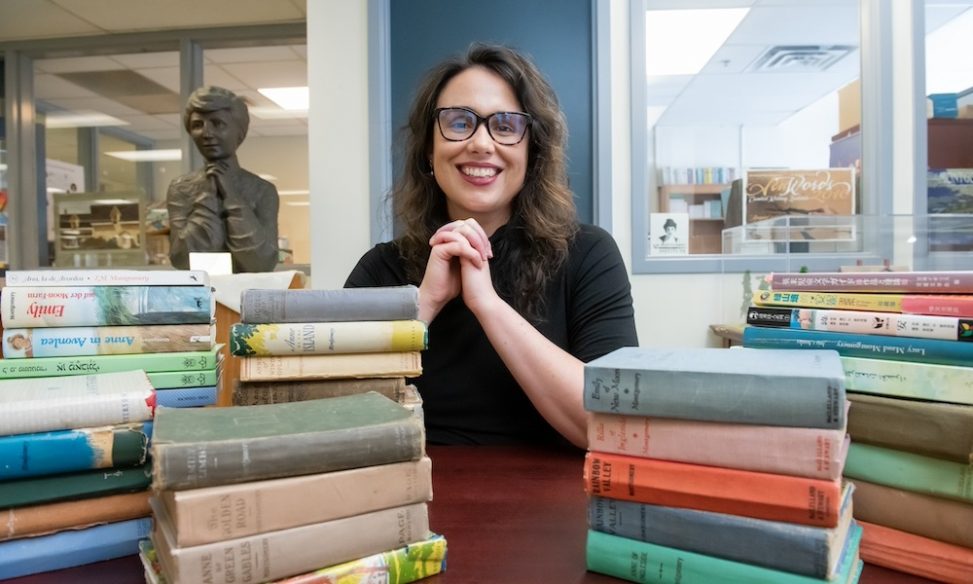 Dr. Kate Scarth sitting in behind stacks of books with a bust of Lucy Maud Montgomery behind left