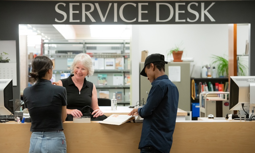 a library worker at the Service Desk helping two students